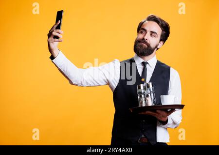 Junger Kellner, der Fotos im Studio macht und mit dem Handy ap dumme Bilder von ihm mit der Platte in der Hand macht. Butler, der im Restaurant arbeitet, nimmt Selfies vor gelbem Hintergrund. Stockfoto