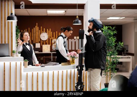 Der junge Mann kommt im luxuriösen Skigebiet an und das freundliche Personal begrüßt ihn in der gemütlichen Hotellobby. Männlicher Touristenkorrespondent für Winteraktivitäten mit Skiausrüstung. Ein perfekter Winterurlaub erwartet Sie. Stockfoto