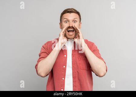 Staunender junger Mann, der mit den Händen um den Mund brüllte und die Kamera auf grauem Hintergrund betrachtete. Stockfoto