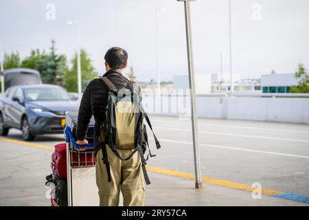 Mann, der den Gepäckwagen schiebt und zu einem geparkten Auto vor dem Flughafen läuft. Stockfoto