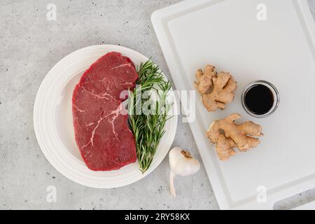 Rohes Rindersteak mit Ingwerwurzel, Knoblauch, Sojasauce und Rosmarin in Nahaufnahme auf dem Küchentisch, flach liegend mit Kopierplatz Stockfoto