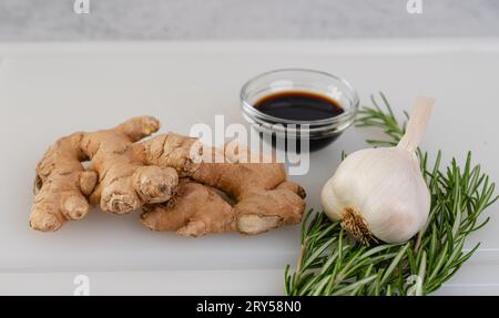 Knoblauch, Rosmarin, Sojasauce und Ingwerwurzel aus nächster Nähe auf einem weißen Schneidebrett auf dem Küchentisch Stockfoto