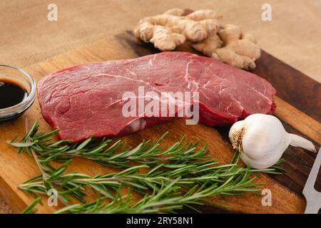 Rohes Rindersteak mit Ingwerwurzel, Knoblauch, Sojasauce und Rosmarin auf einem Holzbrett auf dem Küchentisch Stockfoto