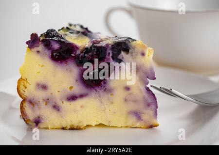 Blick auf die Scheibe Joghurt-Blaubeerkuchen mit Metallgabel in einer weißen Keramikschale und einer Tasse Kaffee. Stockfoto