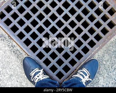 Herrenbeine in Jeans und blaue Ledersneaker in der Nähe des Metallgitters des Sturmabflusses. Lustige Aussicht. Nahaufnahme. Selektiver Fokus. Leerzeichen kopieren. Stockfoto