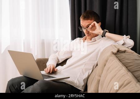 Erwachsener Mann, der empört auf den Laptop-Bildschirm schaut, für den er arbeitet. Geschäftsmann, der zu Hause operiert, auf einer weichen Couch sitzt und überraschend auf die com starrt Stockfoto