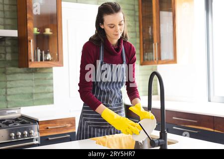 Glückliche kaukasische Frau mit Schürze, die zu Hause in der Küche Geschirr macht Stockfoto