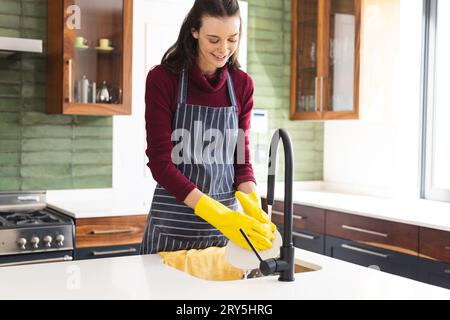Glückliche kaukasische Frau mit Schürze, die zu Hause in der Küche Geschirr macht Stockfoto