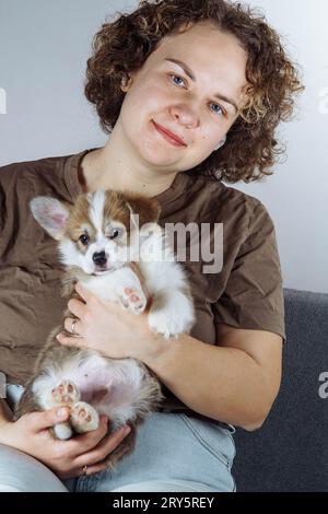 Porträt einer positiven jungen Frau mit lockigen dunklen Haaren in Jeans, braunem T-Shirt mit dem schönen kleinen braunen weißen welpen des Hundes welsh pembroke c Stockfoto