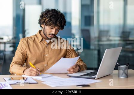 Junger seriöser, fokussierter und denkender Geschäftsmann, Papierarbeit, erfolgreicher hispanischer Mann, der Vertragsberichte und Rechnungen prüft, mit Laptop am Schreibtisch im Büro sitzt, das Formular prüft und ausfüllt. Stockfoto