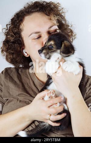 Porträt einer glücklichen jungen Frau mit lockigen Haaren, die ein braunes T-Shirt trägt, das an den Händen hält und den kleinen schwarzen weißen welpen des Hundes welsh pembroke Corgi küsst Stockfoto