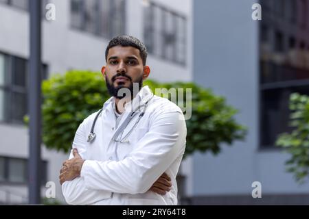 Porträt eines selbstbewussten muslimischen männlichen Arztes, der vor einem Krankenhaus steht und ernsthaft in die Kamera schaut, während seine Hände über die Brust gefaltet sind. Stockfoto