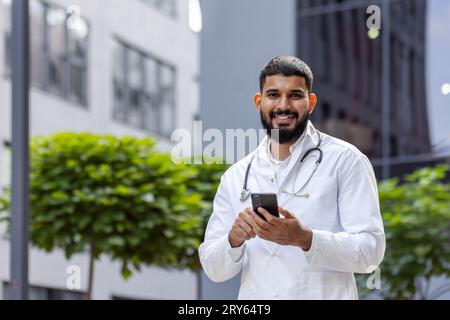 Porträt eines jungen muslimischen männlichen Arztes, der lächelnd in der Nähe der Klinik steht und ein Telefon in den Händen hält. Blick und Lächeln auf die Kamera. Stockfoto