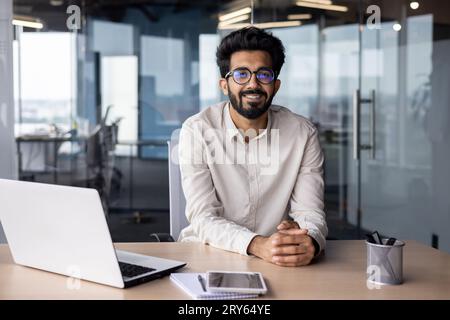 Porträt eines jungen indischen Designers, Ingenieurs, der im Büro am Tisch sitzt und die Kamera betrachtet. Stockfoto