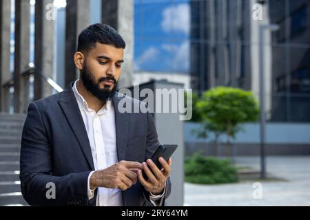 Muslimischer junger Bleistiftgeschäftsmann, der in der Nähe des Bürogebäudes steht und telefoniert, anruft, SMS schreibt, auf ein Treffen wartet. Stockfoto