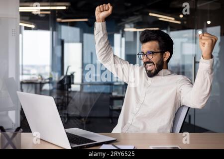 Glücklicher junger indischer Geschäftsmann sitzt im Büro vor einem Laptop und freut sich über den Erfolg, indem er zwei Hände hochhält. Stockfoto