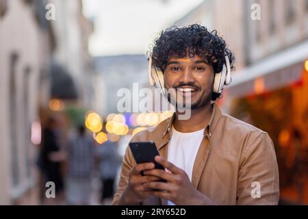 Porträt eines gutaussehenden, lächelnden Indianers, der auf die Kamera schaut. Auf einer Straße in der Stadt mit weißen Kopfhörern stehen und ein Telefon halten. Stockfoto