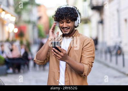 Junger Indianer, der auf der Straße steht, Kopfhörer und Telefon trägt und Musik genießt, lächelnd. Stockfoto