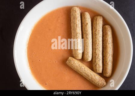 Eine köstliche Gazpacho-Suppe in einer runden Schüssel, garniert mit knusprigen Vollkornbreadsticks, die in einem fast halbkreisförmigen Muster auf einem schwarzen Backgrou angeordnet sind Stockfoto