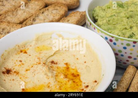 Eine Schüssel Hummus-Dip aus Kichererbsen, Tahini, Knoblauch und Zitrone. Der Hintergrund ist verschwommen, aber man sieht eine Schüssel Guacamole Stockfoto