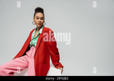 Stilvolles afroamerikanisches Modell in trendigem und lebendigem Outfit, das auf einem Würfel auf grauem Hintergrund sitzt Stockfoto