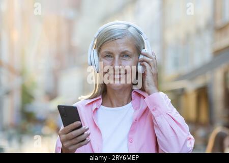 Porträt einer älteren, schönen grauhaarigen Frau, die Kopfhörer trägt und Musik am Telefon hört. Auf einer Straße in der Stadt stehen und die Kamera anlächeln. Nahaufnahme. Stockfoto
