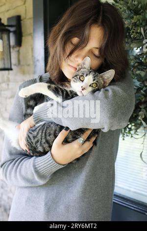 Junge Frau mit einer grau-weißen, tabby-jungen Katze, die sie aus dem Tierheim adoptierte. Selektiver Fokus mit verschwommenem Hintergrund. Stockfoto