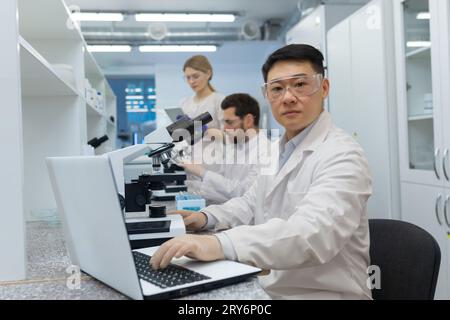 Eine Gruppe junger Studenten von Wissenschaftlern und Chemikern studiert und forscht im Labor der Universität. Asiatischer junger Mann sitzt auf einem Laptop und schaut ernsthaft auf die Kamera. Stockfoto