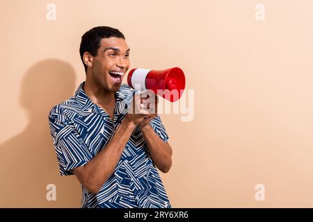 Fotoporträt eines hübschen jungen Mannes, der einen leeren Raum schreit Megaphon trägt trendiges blaues Print-Outfit isoliert auf beigefarbenem Hintergrund Stockfoto