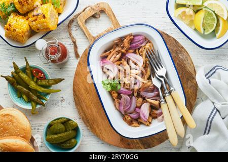 Gezapftes bbq-Hühnchen mit gebackenen Zwiebeln auf Servierplatte, bbq-Mais, Gurken, Chilischoten und Brötchen für Hot Dogs und Burger, Tomatensauce. Traditionell Stockfoto