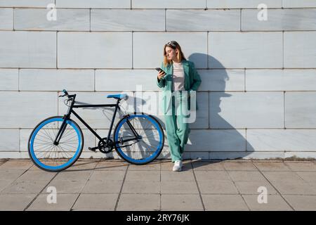 Junge, leistungsfähige, dunkelhaarige Frau im grünen Anzug, die ihr Smartphone mit ihrem Fahrrad auf Marmorhintergrund steht Stockfoto
