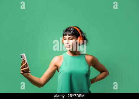 Porträt einer jungen Frau, die Musik auf ihrem Telefon genießt, mit rotem Kopfhörer und grünem Oberteil vor einem passenden grünen Hintergrund. Stockfoto