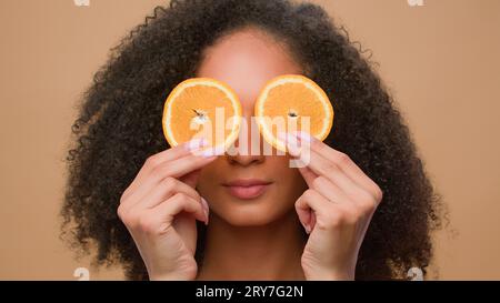 Afroamerikanisches Mädchen, das die Augen mit Zitrusscheiben bedeckt, die die Augen blitzen lassen Frau hält zwei Hälfte der orangen Kosmetik-Dermatologie-Hautpflege-Haarpflege Stockfoto