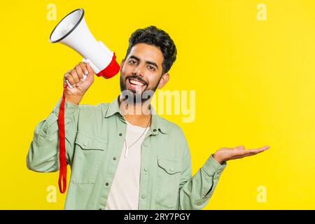 Indischer junger Mann, der mit Megaphon spricht, Nachrichten verkündet, Werbung Rabatte verkauft, Lautsprecher benutzt, um Sprache zu schreien. Glücklicher Araber, isoliert auf gelbem Hintergrund Stockfoto