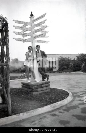 1960er Jahre, historisch, ein junger Mann und eine junge Frau stehen für ein Foto an einem Wegweiser mit mehreren Richtungspfeilen zu den verschiedenen Gegenden in einem Ferienlager in England, Großbritannien. Zu den Orten auf den Schildern gehören: French Coffee Bar, Putting Green, Swimming Pool & Lounge, Theaterspiele & TV Room Lobster Pot Theatre, Wiener Ballsaal, French Bar, Montgomery Ballroom, Regency Theatre. Stockfoto