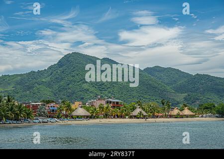 Zihuatanejo, Mexiko - 18. Juli 2023: Playa La Madera mit strohbedeckten, runden, offenen Hütten, die Schatten vor grünen bewaldeten Hügeln bieten Stockfoto