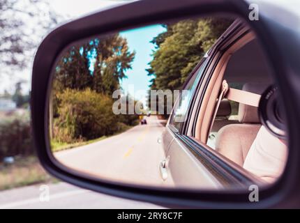 Seitenrückspiegel bei einem Auto. Reflexion der sonnigen Herbststraße am Autoseitenspiegel. Schauen Sie in den Rückspiegel eines Fahrzeugs. Landschaft in der si Stockfoto