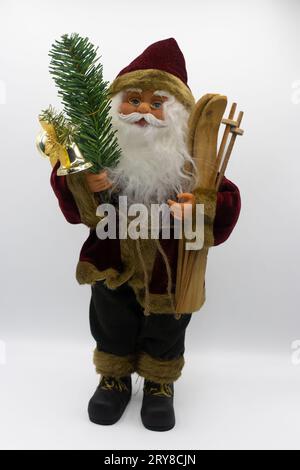 Das Weihnachtsmann-Spielzeug hält Holzskier in der einen Hand und einen Tannenzweig mit einer Glocke in der anderen auf weißem Hintergrund Stockfoto