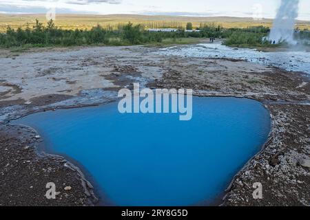 Heiße Quellen, geothermisches Gebiet im Haukadalur Valley, Blesi heiße Quelle vor dem Haus, Strokkur Geysir, der dahinter ausbricht, Golden Circle, Southern Region, Island. Stockfoto