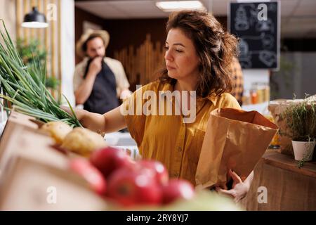 Frau in einem Zero Waste Shop, wo sie Gemüse vom Bauernhof kauft und reife grüne Zwiebeln pflückt. Kunde in einem ortsansässigen, kunststofffreien Lebensmittelgeschäft, in dem eine zersetzbare Papiertüte verwendet wird, um den Klimawandel zu vermeiden Stockfoto