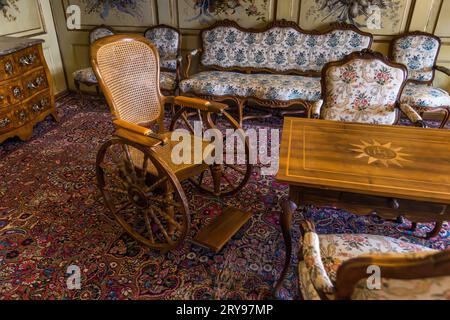 Der antike Holzrollstuhl mit einem Stockersitz und Fußstütze steht im Vordergrund eines Salons mit bemalten Blumenwandpaneelen im Schloss Gruyères in Épagny nahe Gruyères, Kanton Freiburg, Schweiz, neben einem Beistelltisch mit eingelegtem Sternenmuster und Monogramm sowie einer Kommode mit Messingbeschlägen. Die Einrichtungsgegenstände stammen wahrscheinlich aus der Zeit, als die in Genf ansässige Familie Bovy das Schloss ab 1849 als Sommerresidenz nutzte. Das im 13. Jahrhundert erbaute Schloss war bis 1554 der Sitz der Grafen von Gruyère. Seit 1938 gehört es dem Kanton Freiburg und dient als Stockfoto