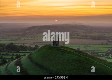 Wanderer, die neben dem St. Michael’s Tower auf dem Glastonbury Tor in Glastonbury in Somerset, Großbritannien, stehen, beobachten den spektakulären Sonnenaufgang im Herbst. Stockfoto