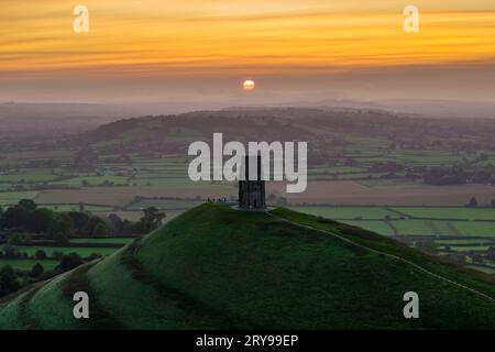 Wanderer, die neben dem St. Michael’s Tower auf dem Glastonbury Tor in Glastonbury in Somerset, Großbritannien, stehen, beobachten den spektakulären Sonnenaufgang im Herbst. Stockfoto