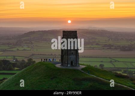 Wanderer, die neben dem St. Michael’s Tower auf dem Glastonbury Tor in Glastonbury in Somerset, Großbritannien, stehen, beobachten den spektakulären Sonnenaufgang im Herbst. Stockfoto