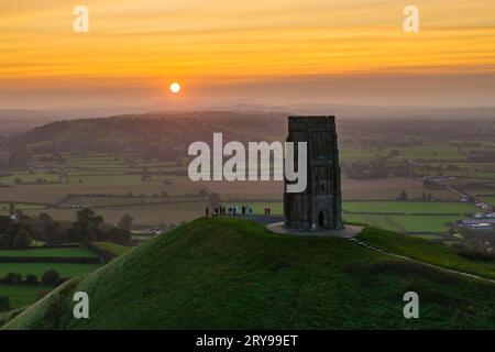 Wanderer, die neben dem St. Michael’s Tower auf dem Glastonbury Tor in Glastonbury in Somerset, Großbritannien, stehen, beobachten den spektakulären Sonnenaufgang im Herbst. Stockfoto