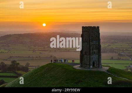 Wanderer, die neben dem St. Michael’s Tower auf dem Glastonbury Tor in Glastonbury in Somerset, Großbritannien, stehen, beobachten den spektakulären Sonnenaufgang im Herbst. Stockfoto