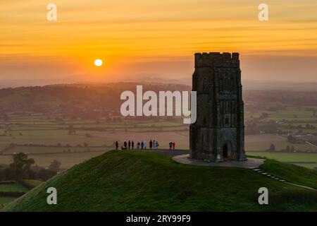 Wanderer, die neben dem St. Michael’s Tower auf dem Glastonbury Tor in Glastonbury in Somerset, Großbritannien, stehen, beobachten den spektakulären Sonnenaufgang im Herbst. Stockfoto