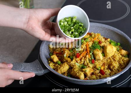 Frau fügt geschnittene grüne Zwiebeln zu Reis mit Fleisch und Gemüse in der Pfanne hinzu, Nahaufnahme Stockfoto