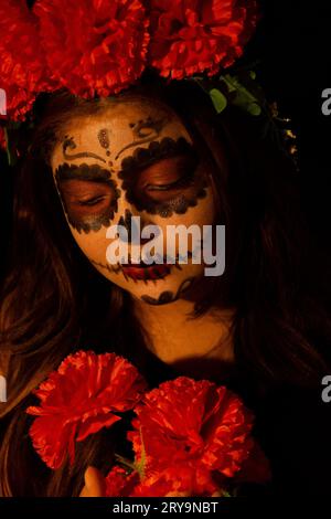 Eine Frau, die als catrina gekleidet ist, auf einem dunklen Hintergrund und einen Blumenstrauß ansieht, den sie in ihren Händen hält Stockfoto
