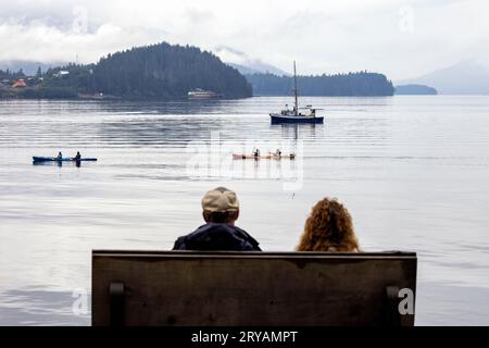 Ein Paar, das auf einer Bank sitzt und Kajakfahrer in Hoonah, Alaska, USA, beobachtet Stockfoto